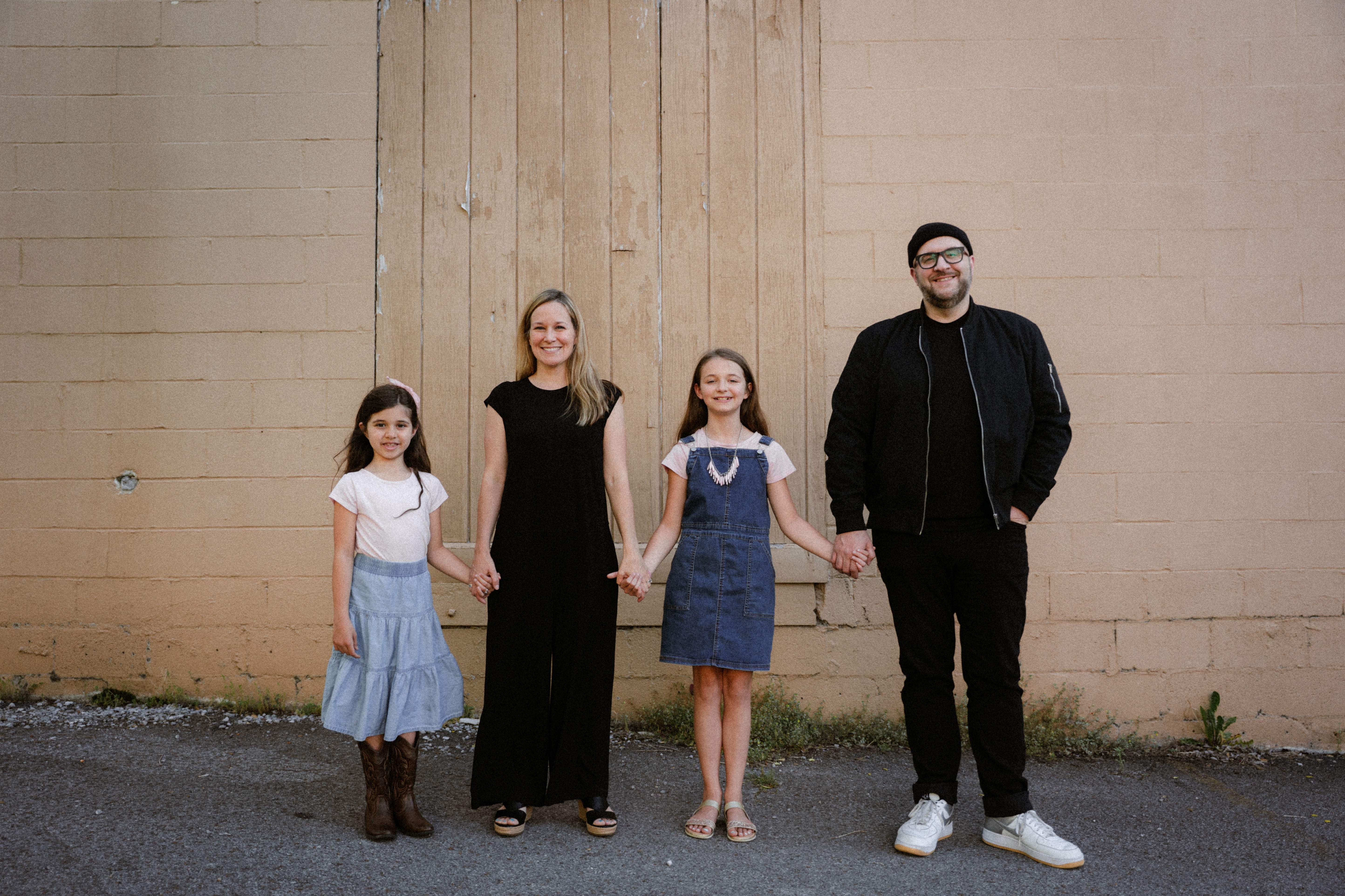 Justin and Naomi Rizzo with their two daughters, holding hands and smiling against a warm textured wall